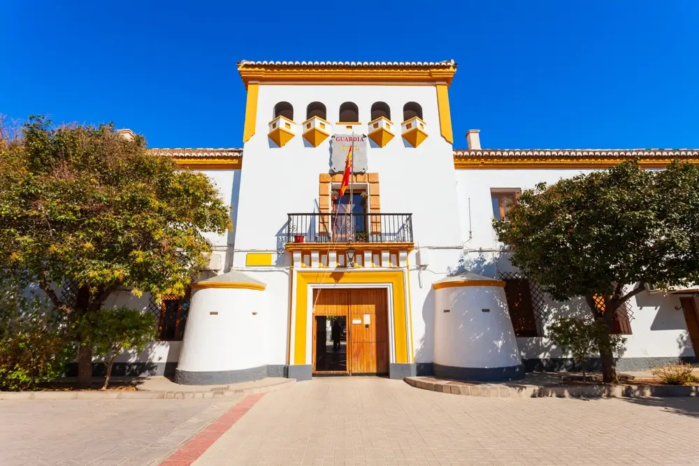 Fachada de un cuartel de la Guardia Civil con la bandera de España y detalles amarillos bajo un cielo azul despejado.