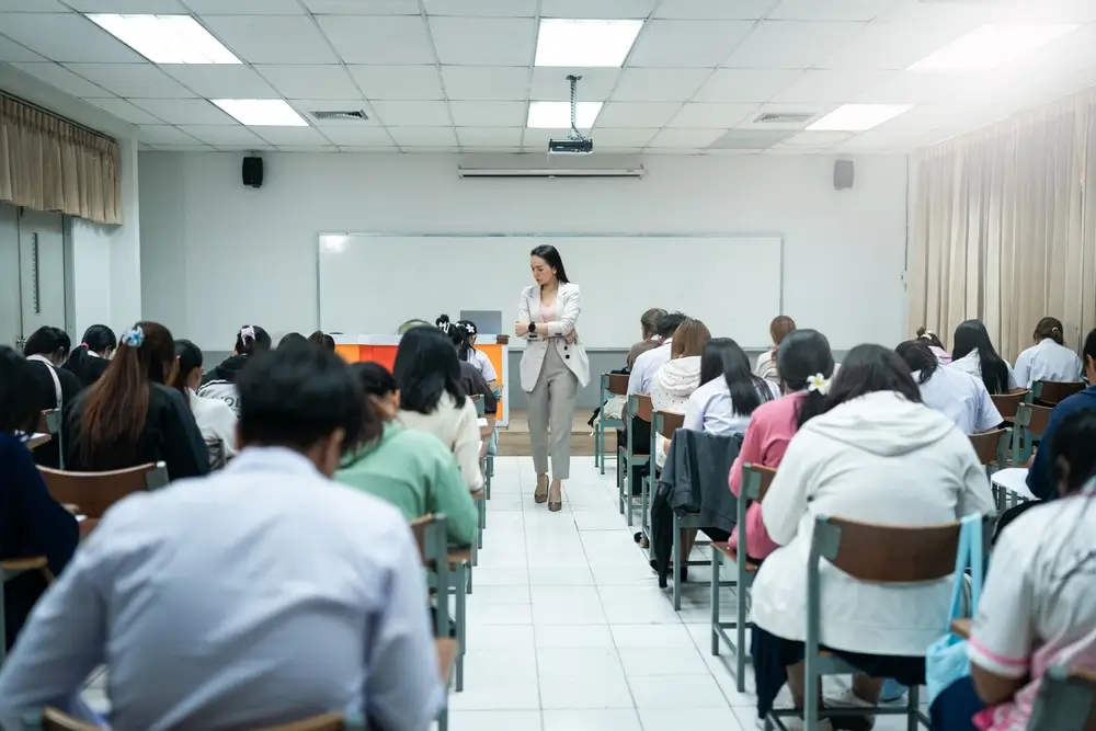 Aspirantes realizando un examen tipo test en un aula de oposiciones mientras una examinadora supervisa la prueba.