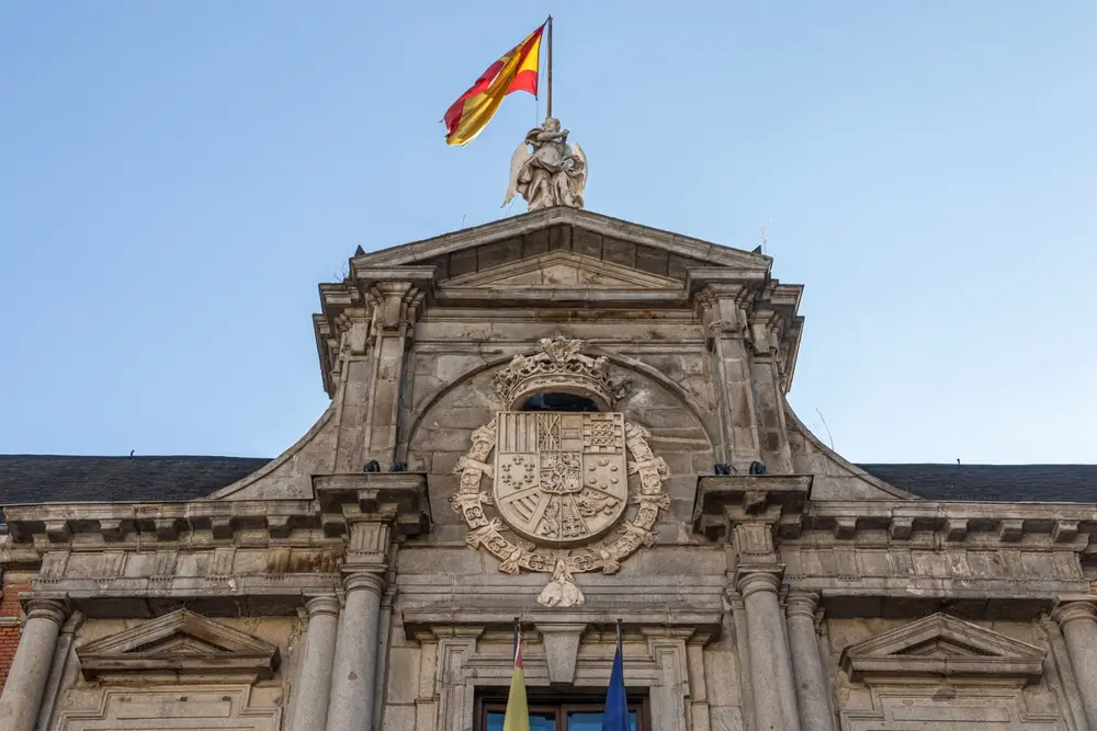 Fachada de la sede de un ministerio en España con escudo institucional de piedra y la bandera nacional en lo alto.