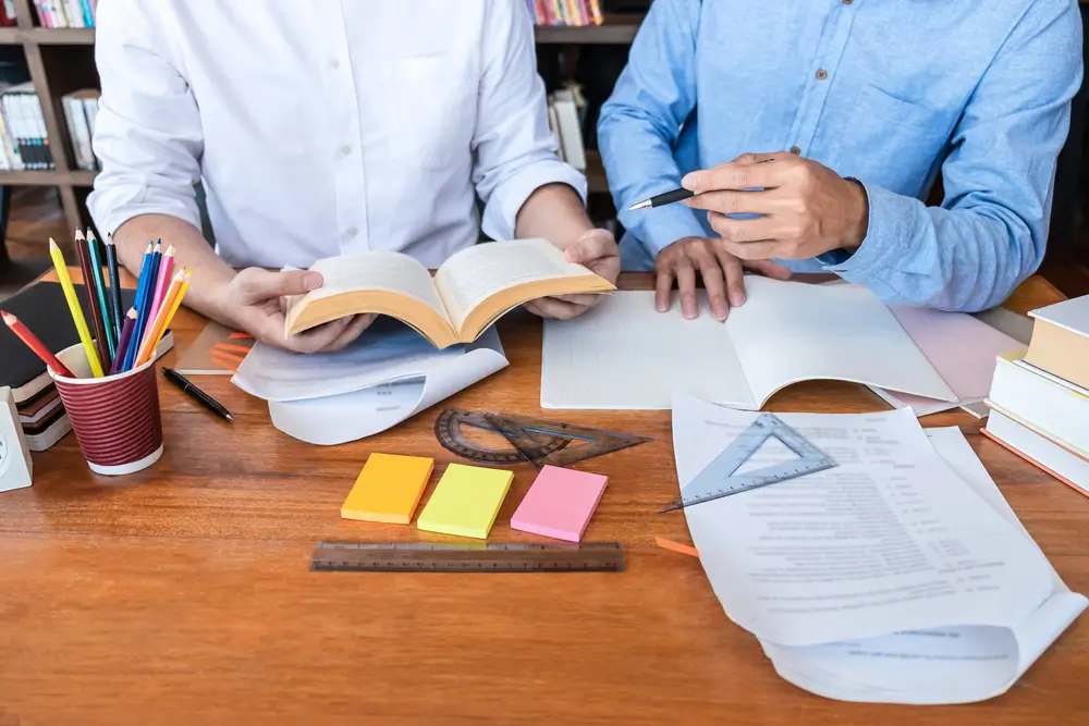 Dos personas consultando libros de estudio y cuadernos sobre un escritorio con material para preparar un examen.