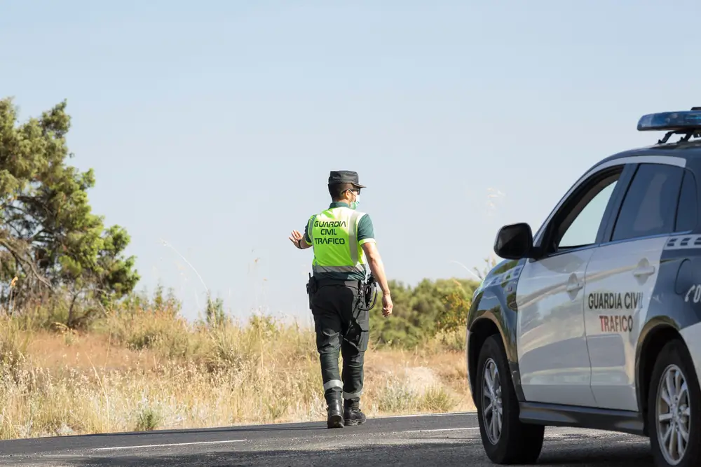 Agente de Tráfico con el uniforme de la Guardia Civil y equipamiento oficial junto a un coche patrulla en carretera.