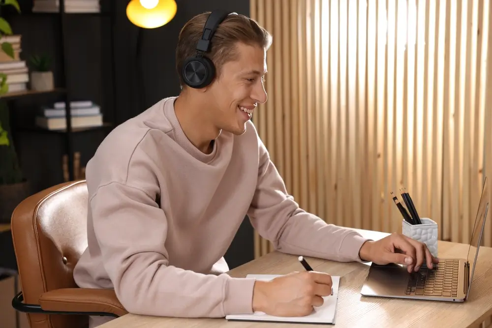 Estudiante sonriente con auriculares preparando un examen con su ordenador portátil mientras toma notas.